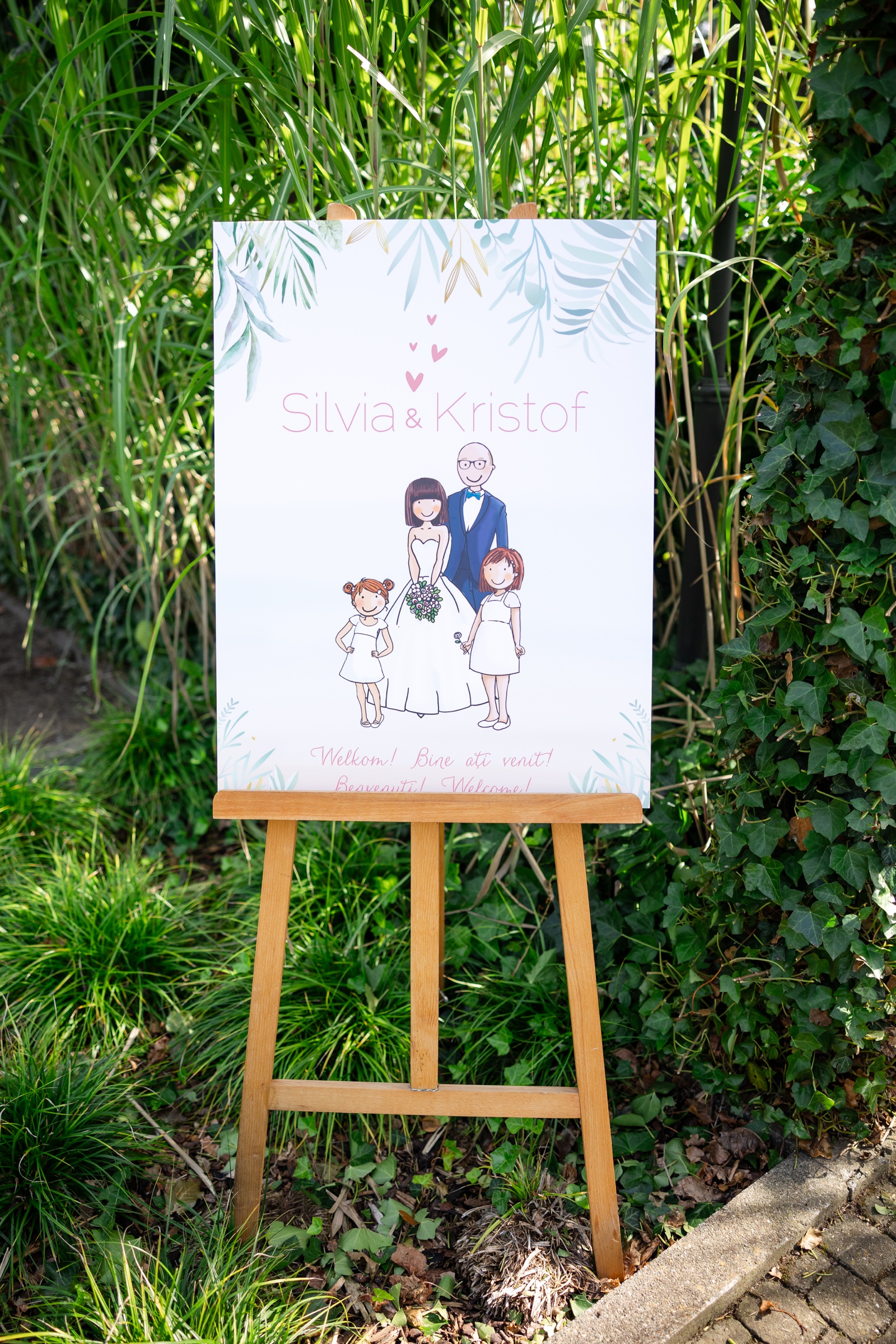 Wedding welcome sign with an illustration of the bride and groom and their two daughters, on a wooden easel.